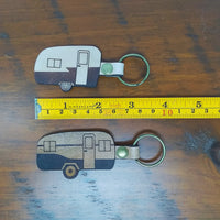 Two keychains shaped like camper trailers on a wooden surface with a measuring tape for scale.