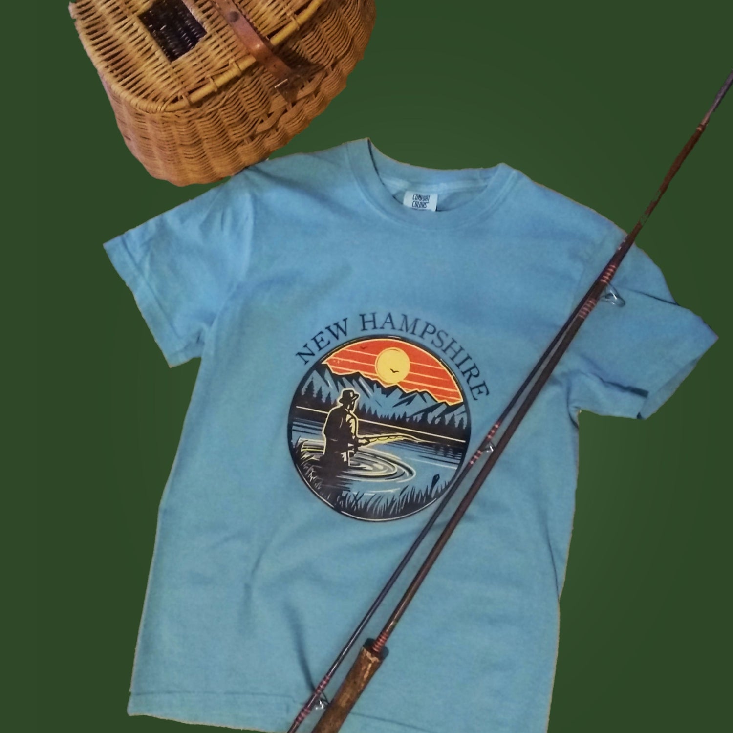 Light blue t-shirt with New Hampshire graphic design, fishing rod, and wicker basket on a green background