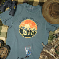 Faded green t-shirt with New Hampshire graphic design on a checkered fabric background with binoculars, hat, and boots.
