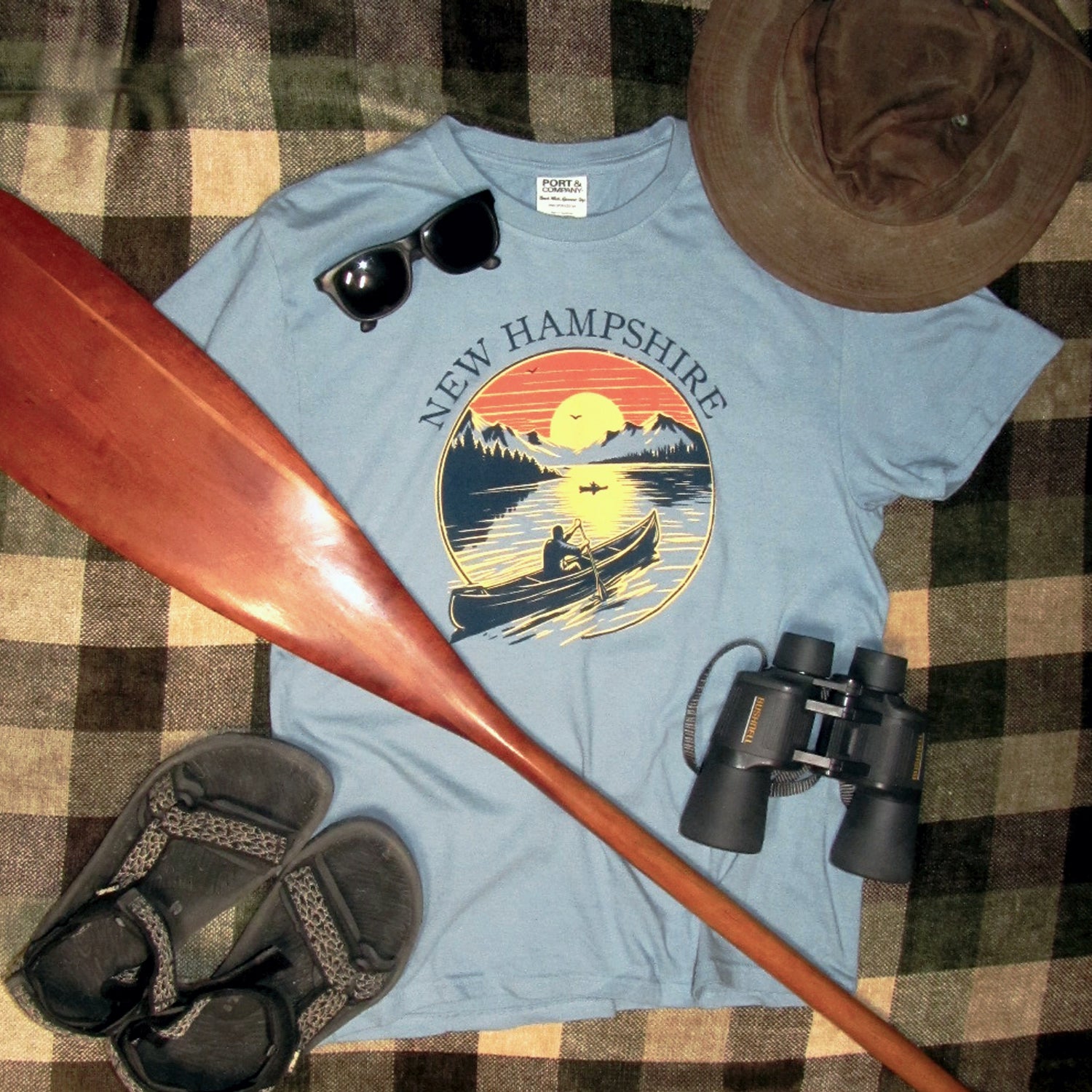 Blue t-shirt with New Hampshire design on a checkered surface with paddle, sunglasses, and binoculars.