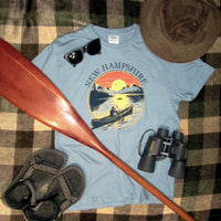 Blue t-shirt with New Hampshire design on a checkered surface with paddle, sunglasses, and binoculars.