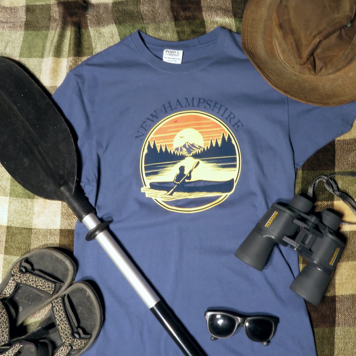 Blue t-shirt with New Hampshire design, paddle, binoculars, sandals, and sunglasses on a checkered background