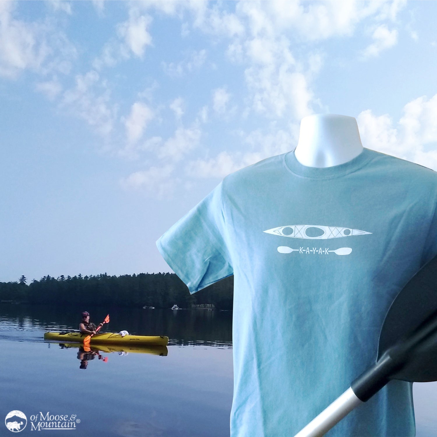 Mannequin wearing a light blue t-shirt with a kayak design against a scenic lake background