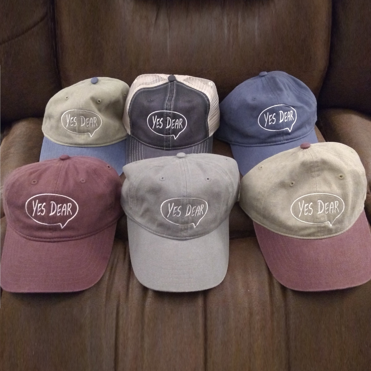 Six baseball caps with 'Yes Dear' text on a brown leather surface