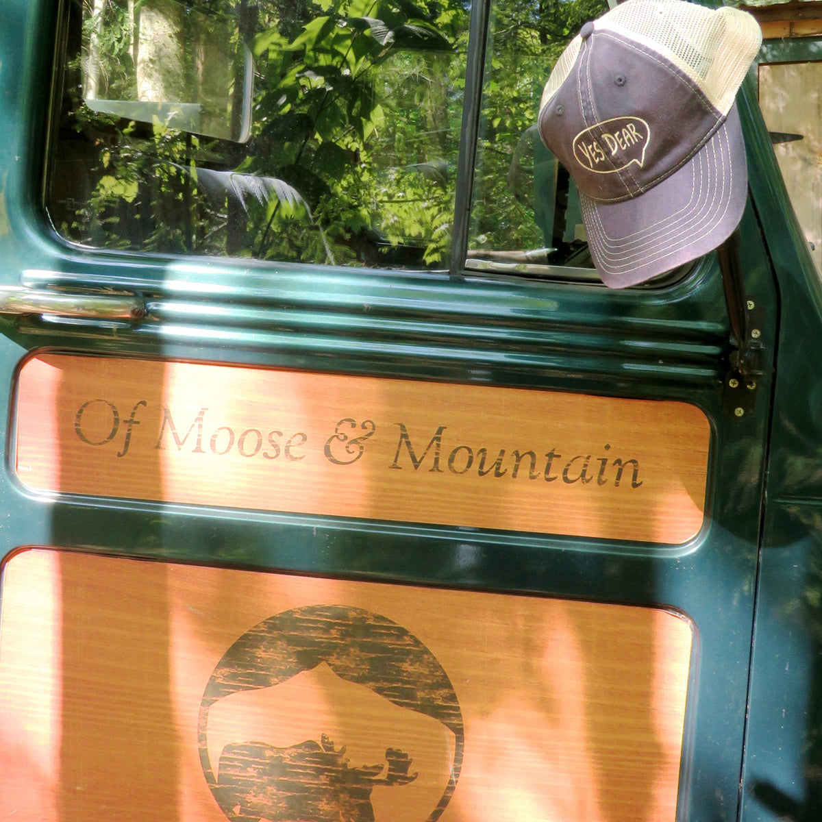 Green vehicle door with 'Of Moose & Mountain' engraving and a Yes Dear cap on the window, surrounded by greenery.
