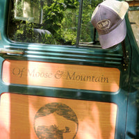 Green vehicle door with 'Of Moose & Mountain' engraving and a Yes Dear cap on the window, surrounded by greenery.