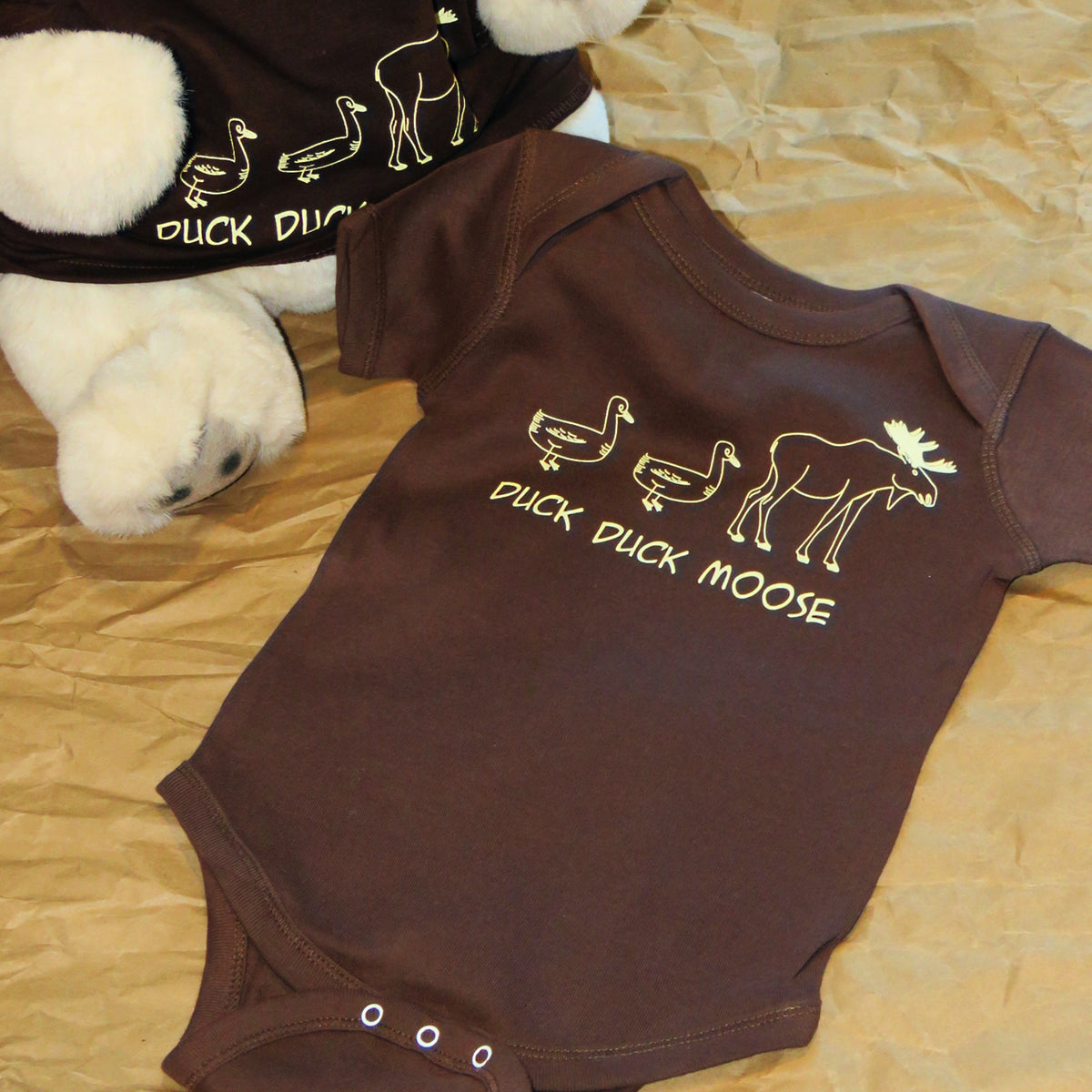 Brown baby onesie with 'Duck Duck Moose' text and graphics on a beige surface.