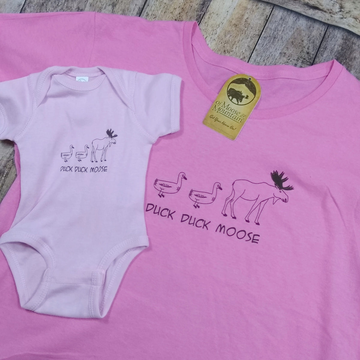 Pink t-shirt and baby onesie with 'Duck Duck Moose' text and graphics on a wooden surface.