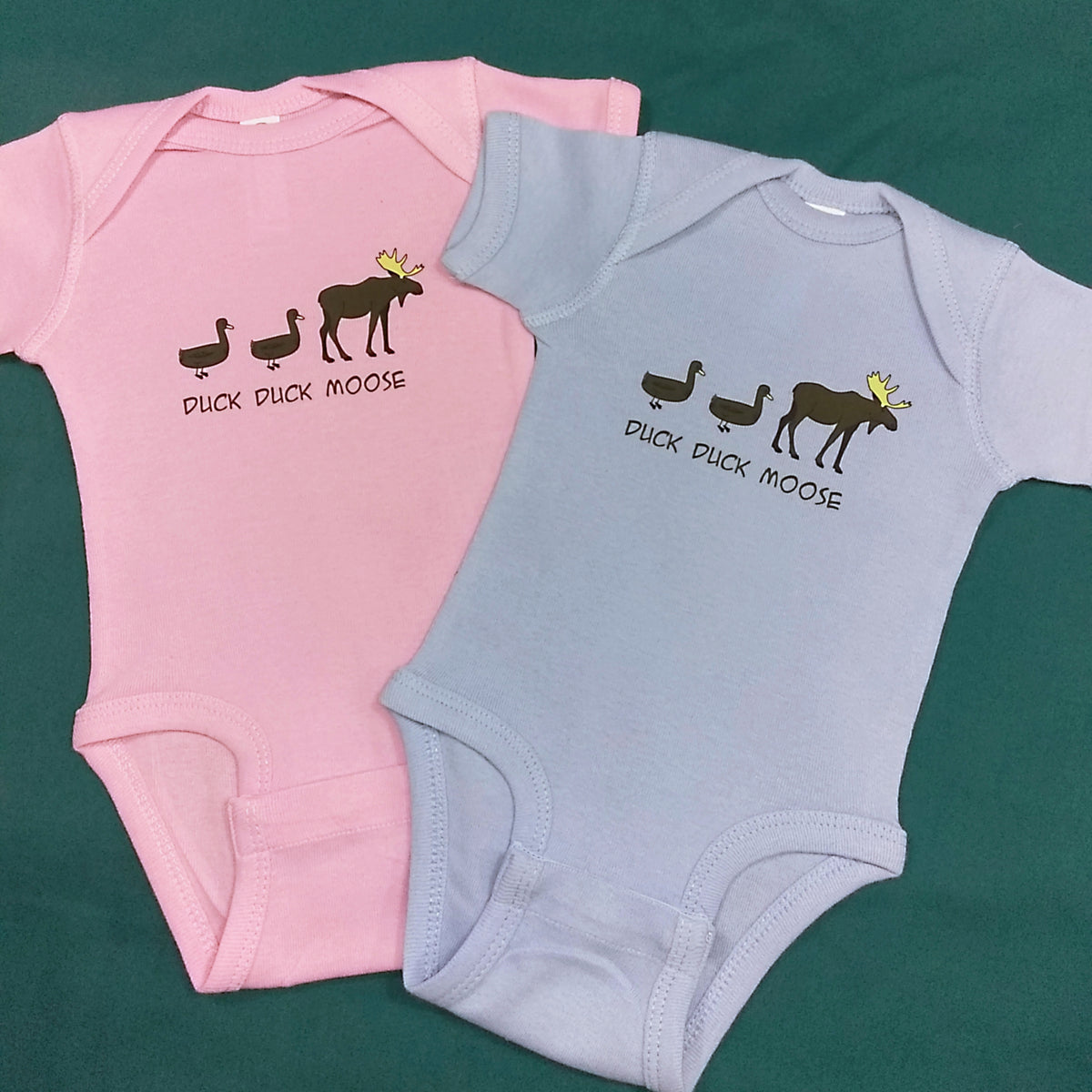 Pink and light blue baby onesies with 'Duck Duck Moose' text and graphics on a green background