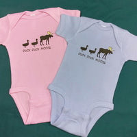 Pink and light blue baby onesies with 'Duck Duck Moose' text and graphics on a green background
