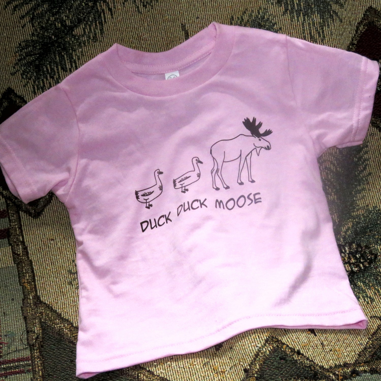Pink t-shirt with 'Duck Duck Moose' text and graphics on a textured surface