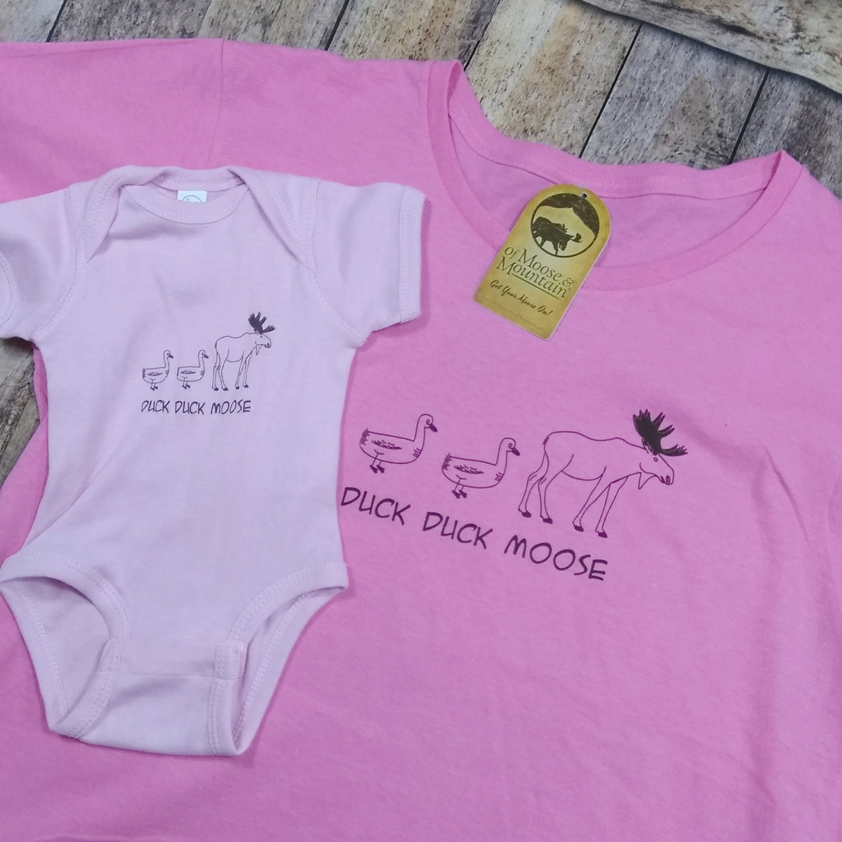 Pink t-shirt and baby onesie with 'Duck Duck Moose' design on a wooden surface