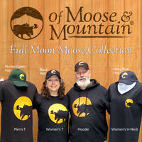 Two people and two mannequins wearing clothing from the 'Full Moon Moose Collection' by of Moose & Mountain against a wooden background.