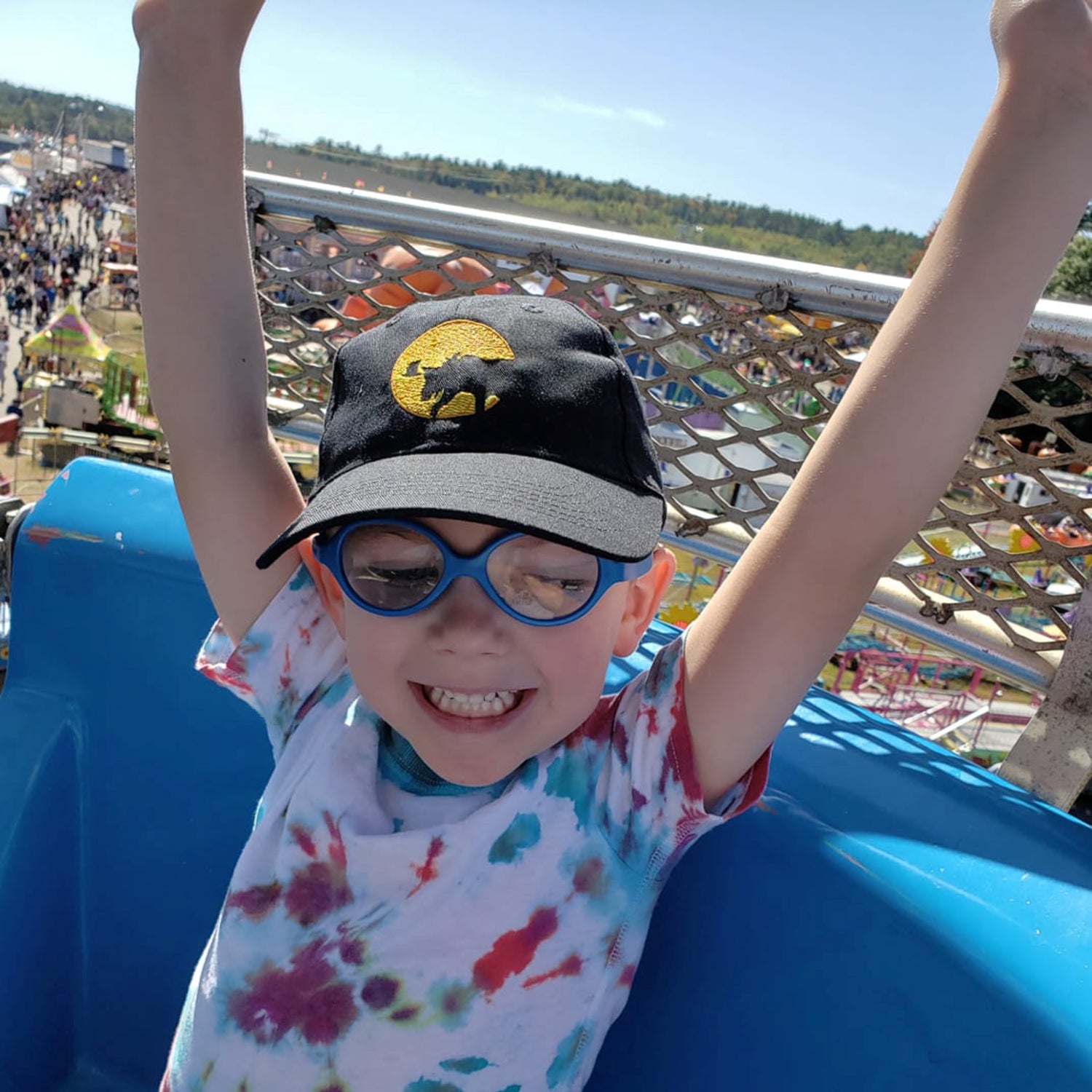 Child with arms raised at an amusement park, wearing a Full Moon Moose cap and colorful shirt.