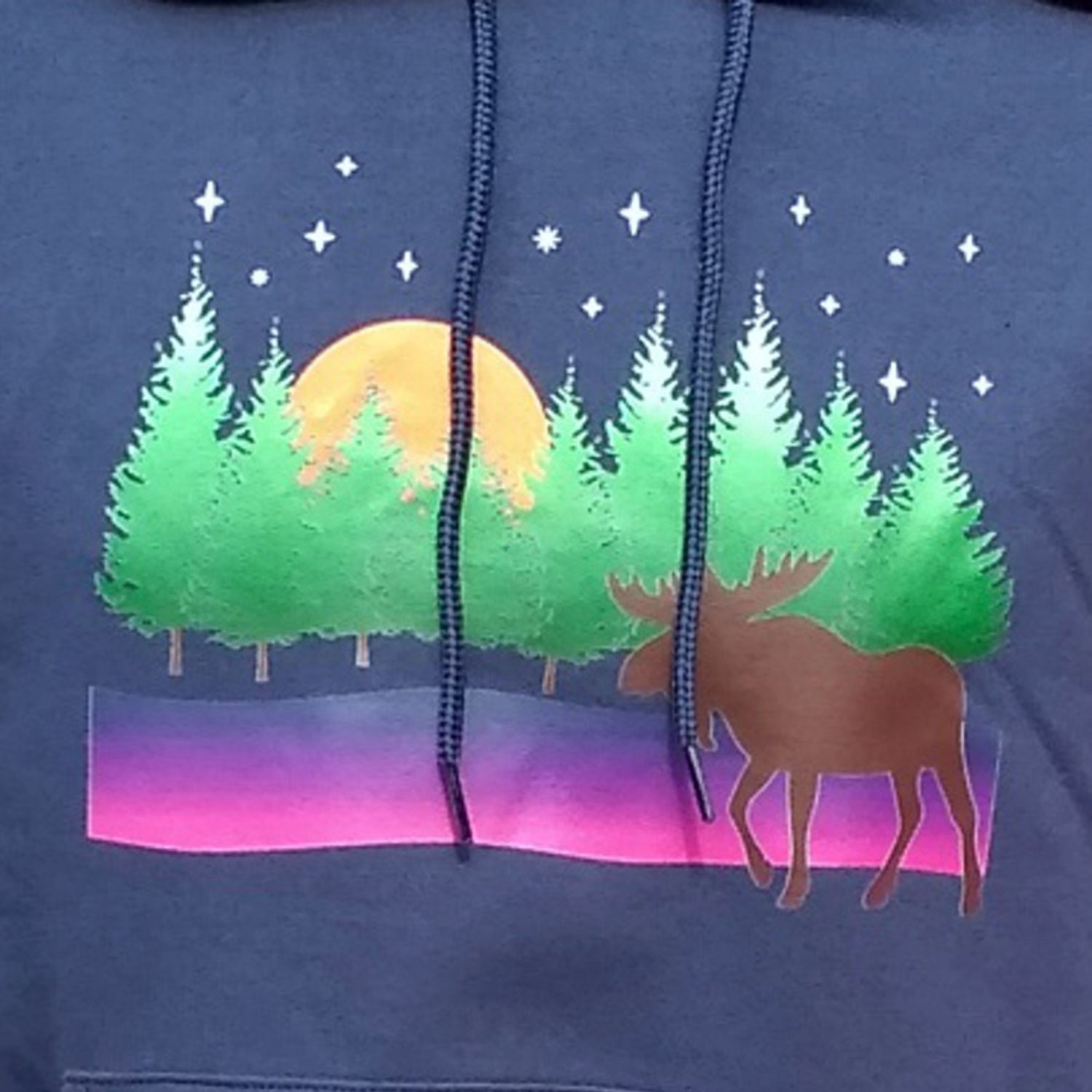 A shirt design featuring a moose in front of a row of trees as the moon rises on a stary night.