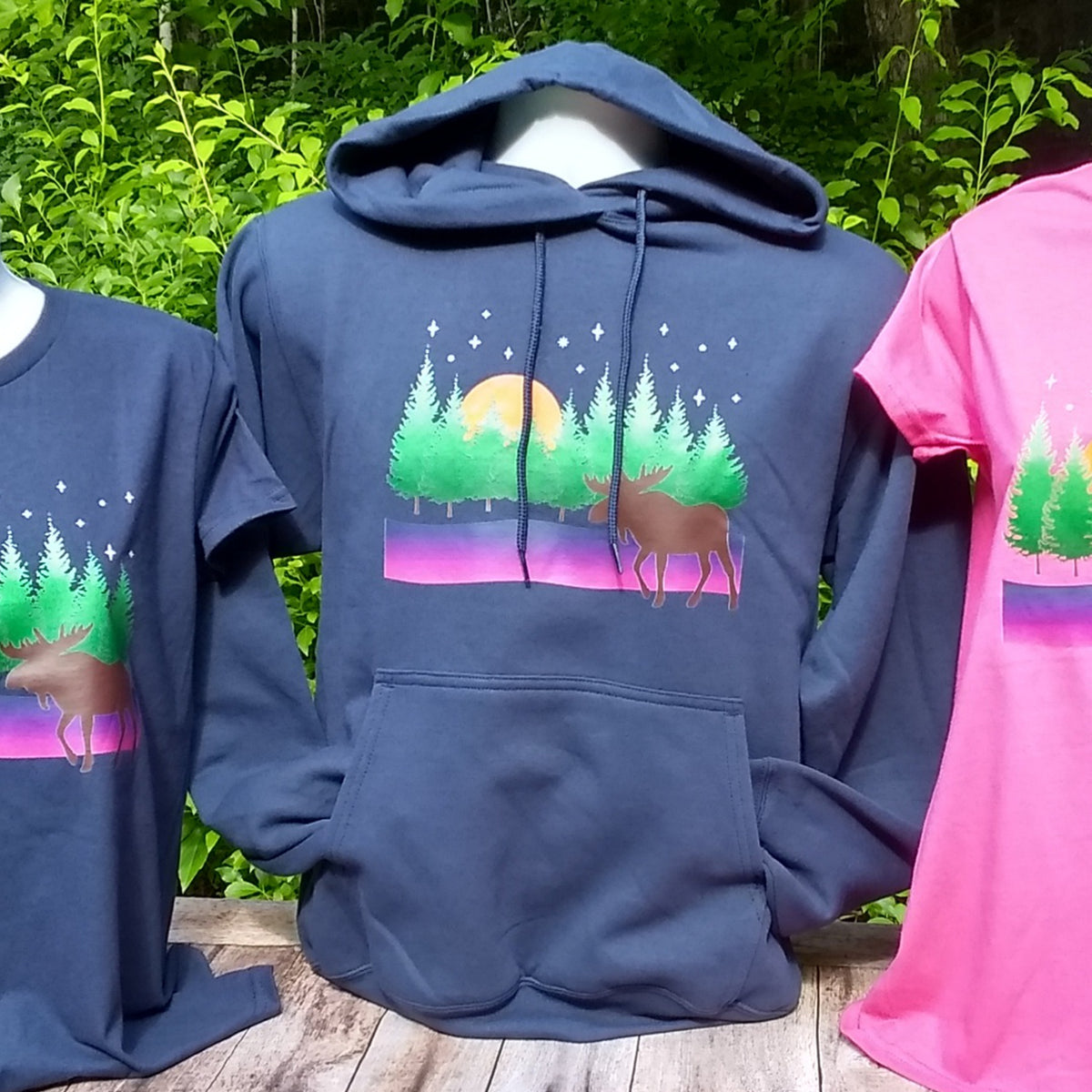 Blue hoodie with moose and forest design on a wooden surface with greenery in the background