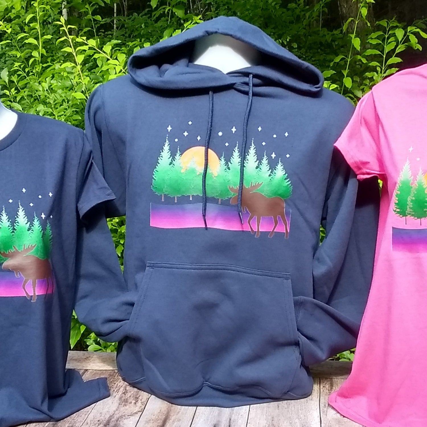 Blue hoodie with moose and forest design on a wooden surface with greenery in the background