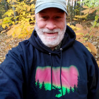 Man wearing a dark hoodie with a moose and forest and Northern Lights design in an autumn forest setting