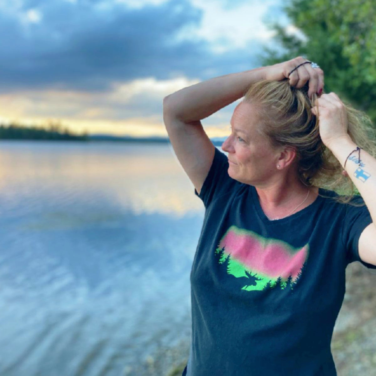 Person standing by a lake with a scenic background wearing a t-shirt with a moose silhouette in front of the northern lights