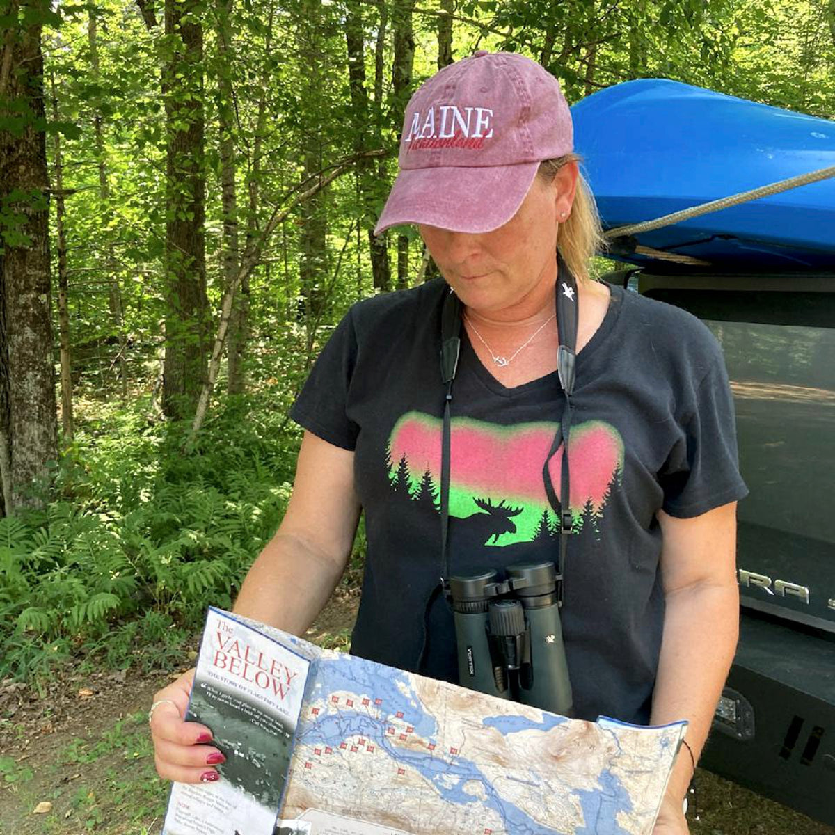 Person in a forest holding a map and wearing a cap with 'Maine' on it and a t-shirt with a moose silhouette in front of the northern lights.