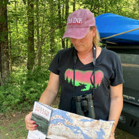 Person in a forest holding a map and wearing a cap with 'Maine' on it and a t-shirt with a moose silhouette in front of the northern lights.