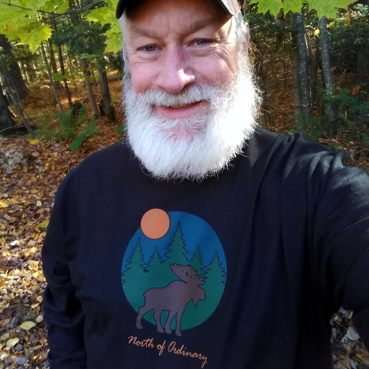Man with a beard wearing a black shirt with a moose design in a forest setting