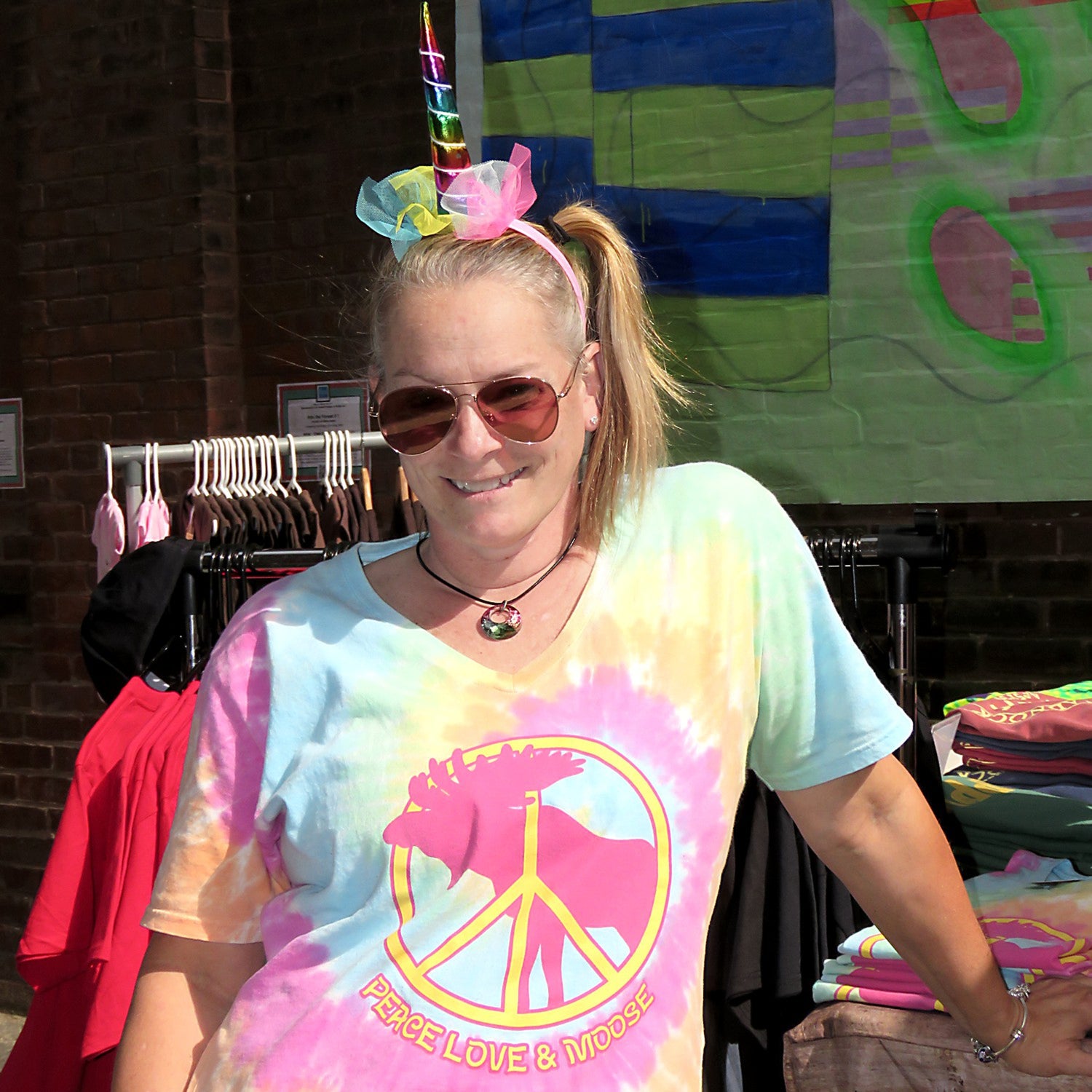 Person wearing a tie-dye shirt with a peace sign and moose design, standing in front of a colorful backdrop.