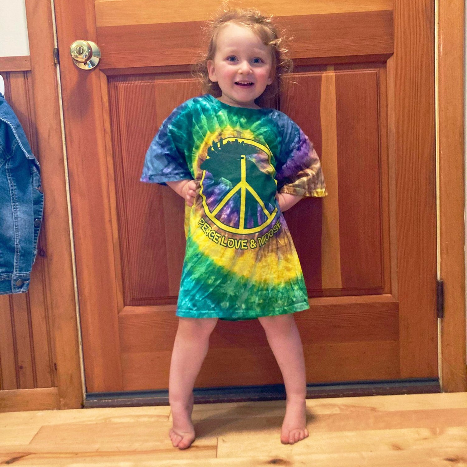 Child wearing a tie-dye shirt with a peace symbol design in front of a wooden door.