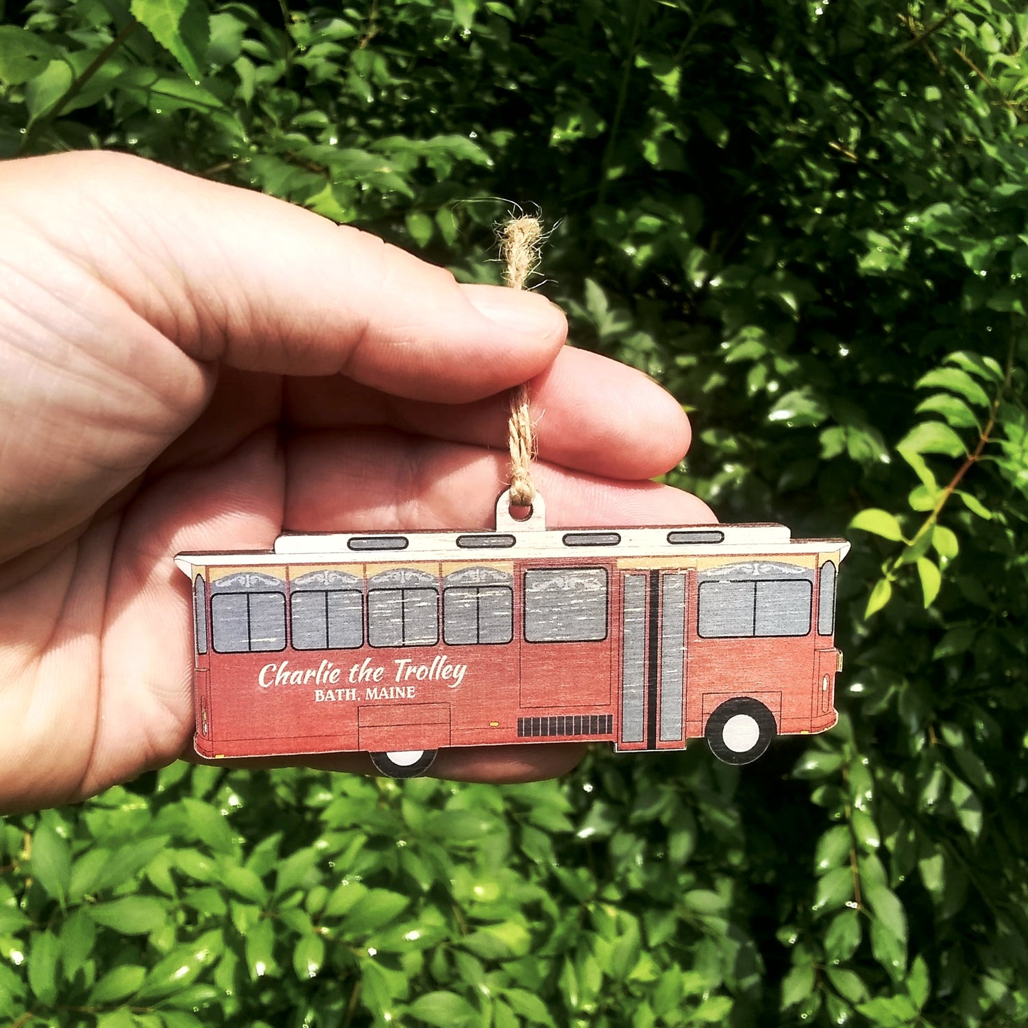 Hand holding a red trolley ornament with 'Charlie the Trolley Bath, Maine' text against a green leafy background.