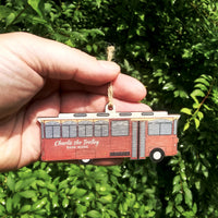 Hand holding a red trolley ornament with 'Charlie the Trolley Bath, Maine' text against a green leafy background.