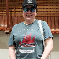 Person wearing a t-shirt with a ship design and text, standing in front of wooden canoe hanging on a wall at a museum.