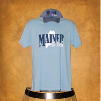 Light blue t-shirt with 'Mainer In Exile' text and a cap on a wooden surface.