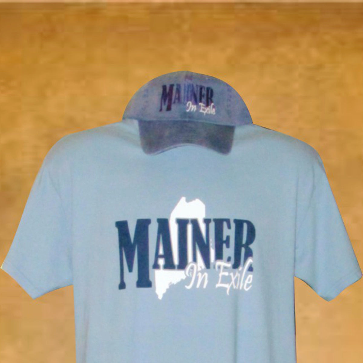 Light blue t-shirt with 'MAINER In Exile' text and a cap on a wooden surface