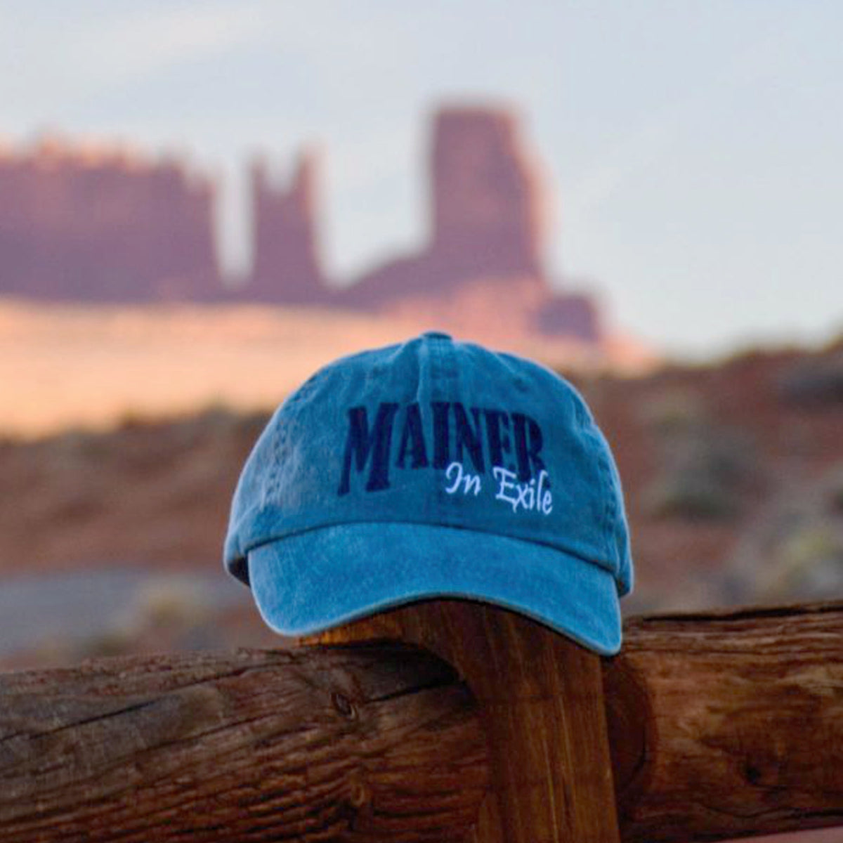 Blue cap with 'MAINE In Exile' text on a wooden post with desert landscape in the background