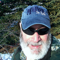 Man wearing a blue cap with text and sunglasses, standing in a snowy forest.