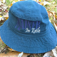 Blue bucket hat with 'Mainer In Exile' text on grass and leaves