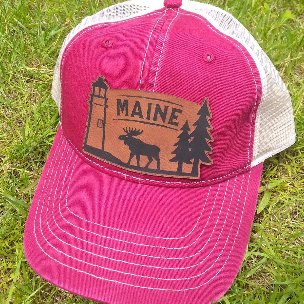 Red cap with 'MAINE' and moose, lighthouse and pine tree design on grass