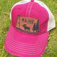 Red cap with 'MAINE' and moose, lighthouse and pine tree design on grass