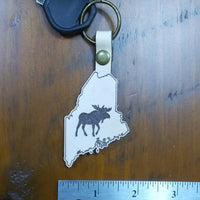 Keychain with Maine shape and moose design on a wooden surface with a ruler for scale.