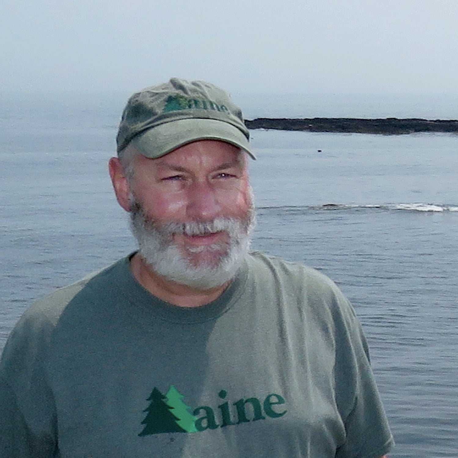 Man wearing a 'Maine' t-shirt and hat standing near the ocean