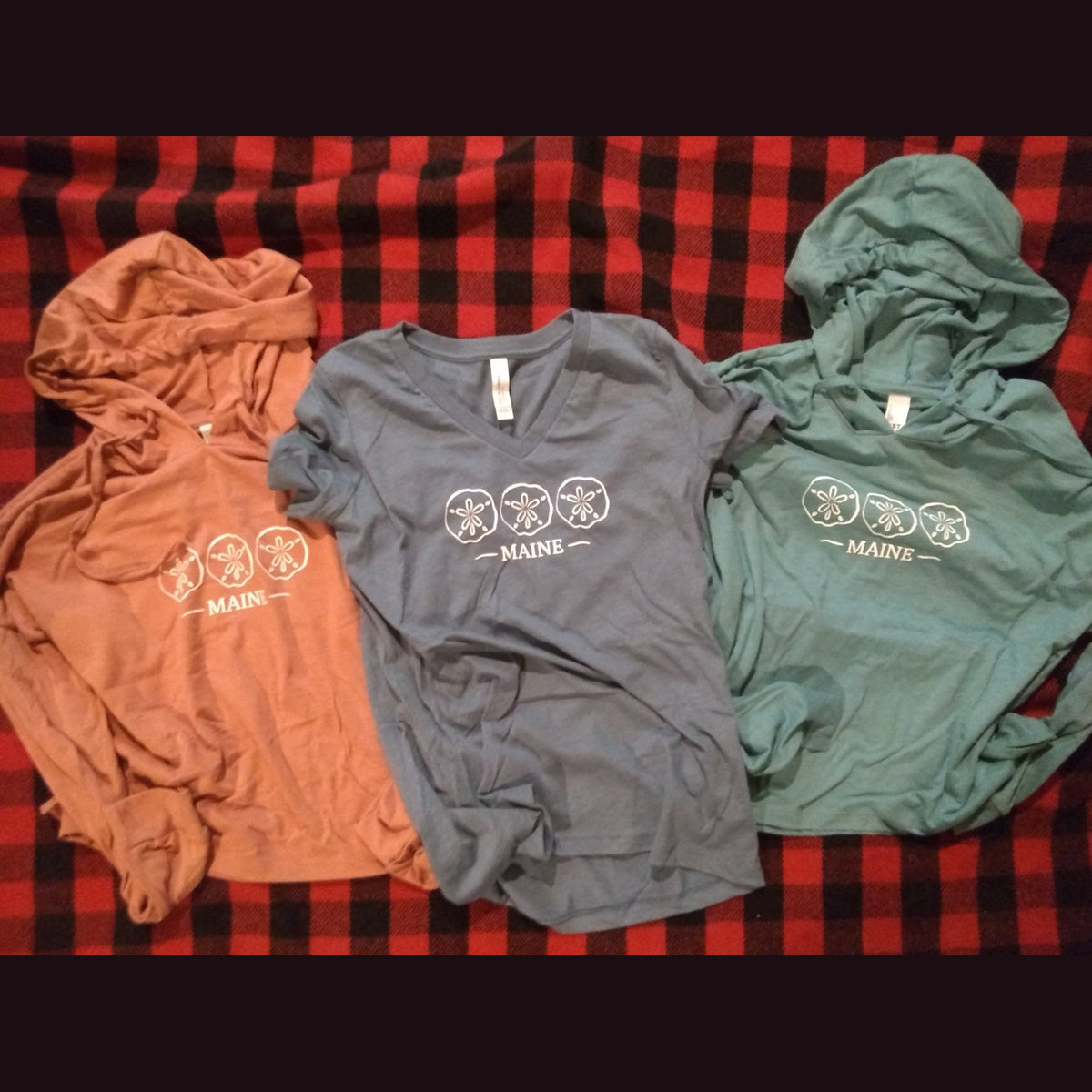 Two hooded t-shirts and a v-neck t-shirt with Maine Sand Dollar design on plaid blanket.
