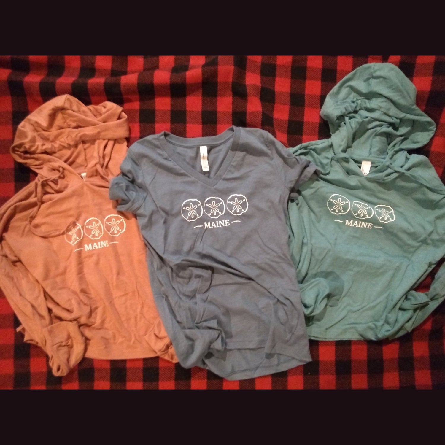 Two hooded t-shirts and a v-neck t-shirt with Maine Sand Dollar design on plaid blanket.