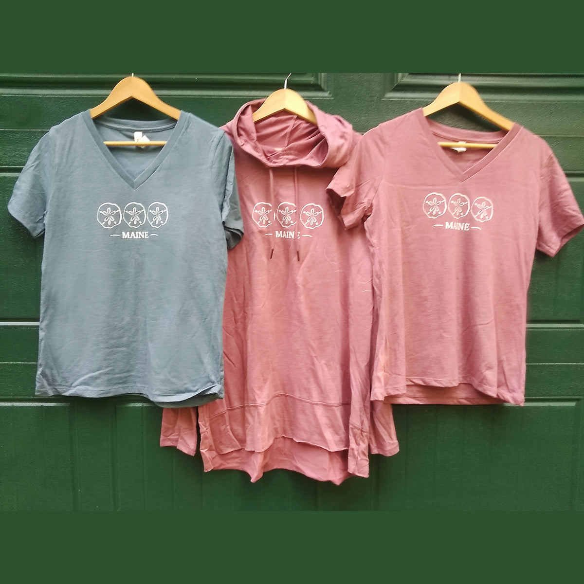 Two pink t-shirts and a gray t-shirt with 'Maine' design on hangers against a green background
