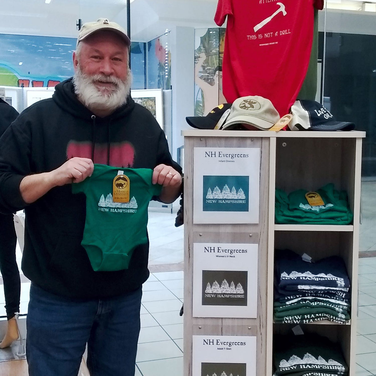 Man holding a green onesie with a logo in front of a display of t-shirts and hats.