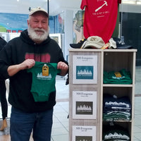Man holding a green onesie with a logo in front of a display of t-shirts and hats.