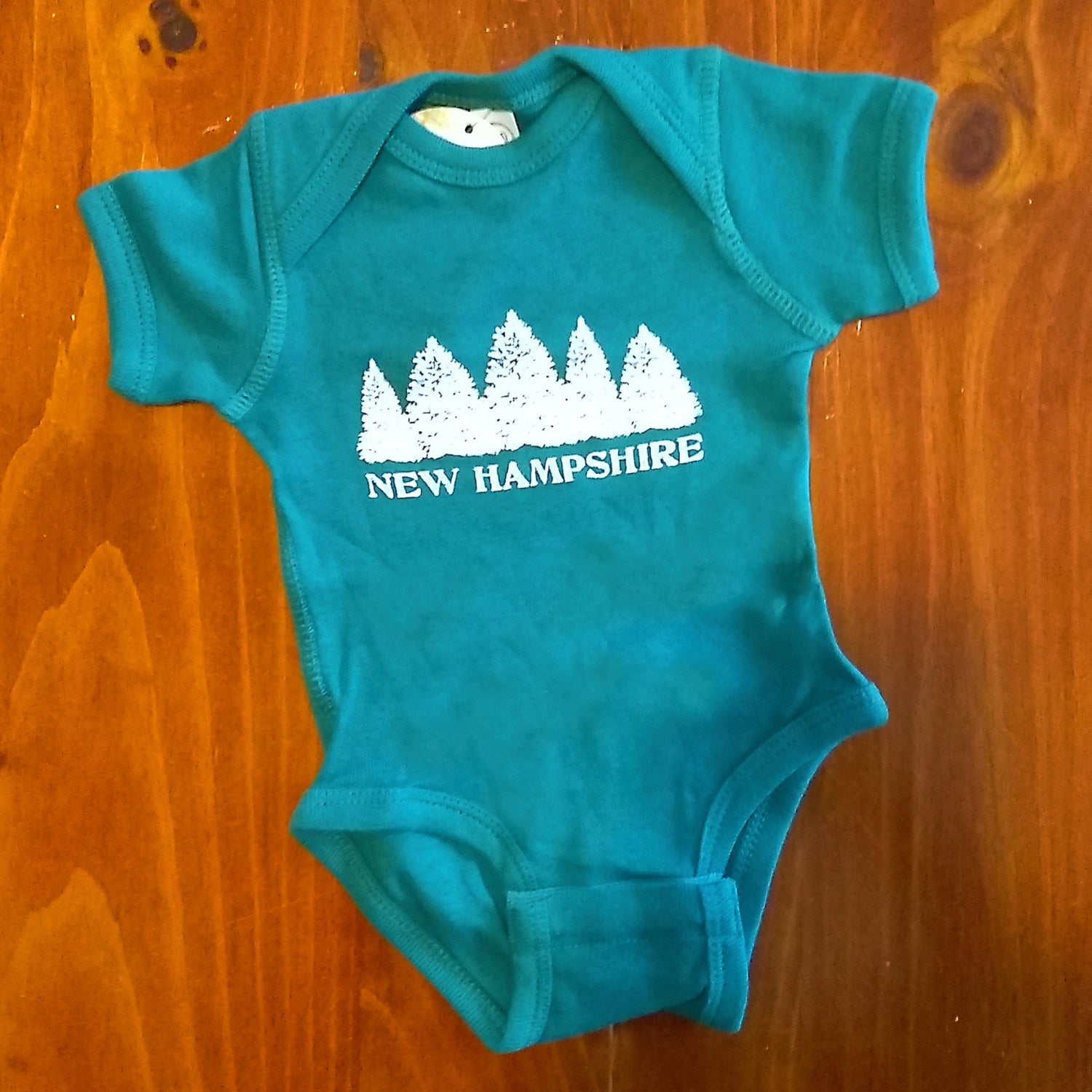 Green baby onesie with 'New Hampshire' and forest design on a wooden surface