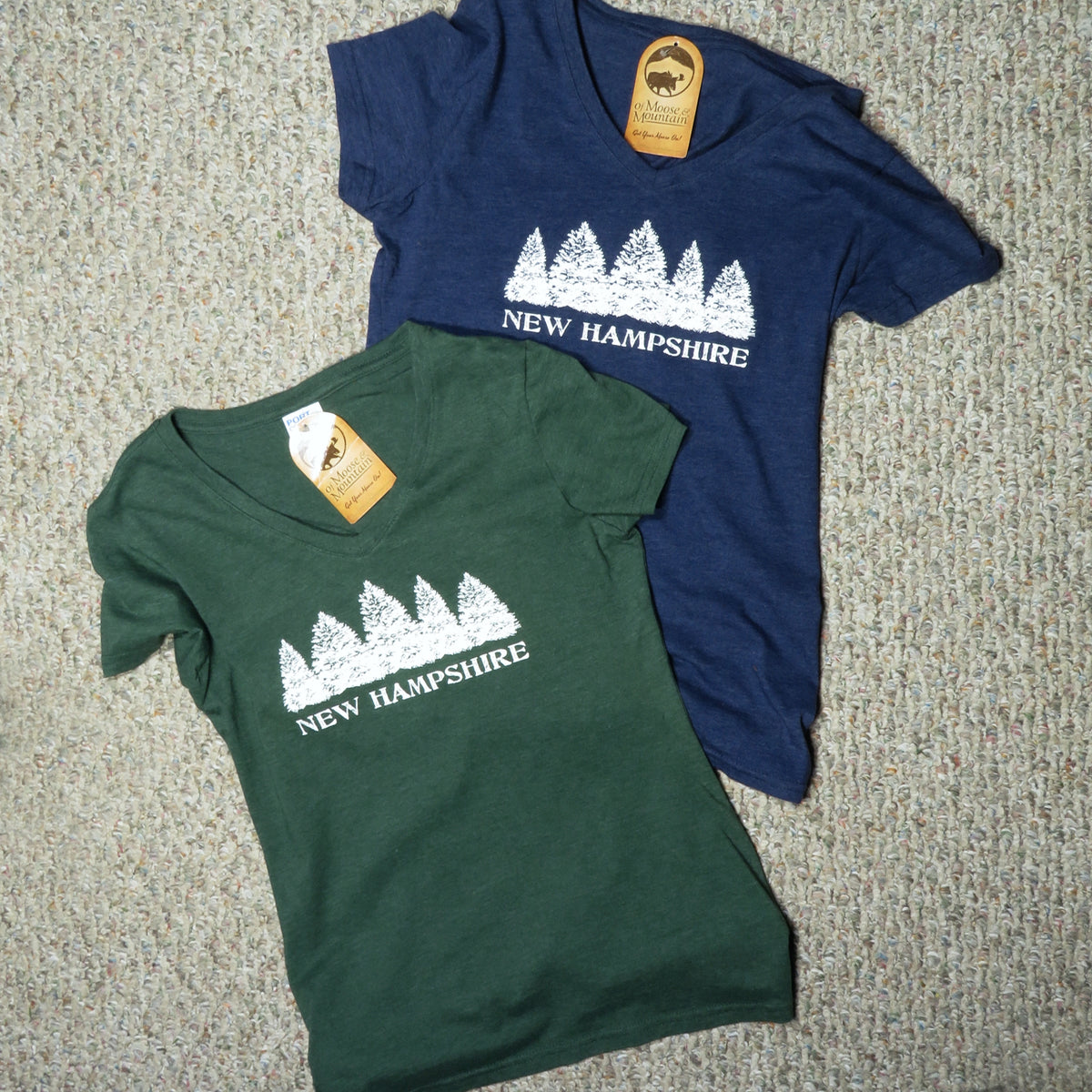 Two t-shirts, one green and one blue, with 'New Hampshire' and a row of evergreen trees printed on them, on a textured surface.