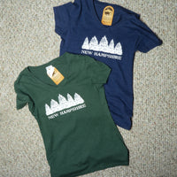 Two t-shirts, one green and one blue, with 'New Hampshire' and a row of evergreen trees printed on them, on a textured surface.