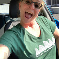 Person wearing a green 'New Hampshire' t-shirt with a row of evergreen trees inside a vehicle