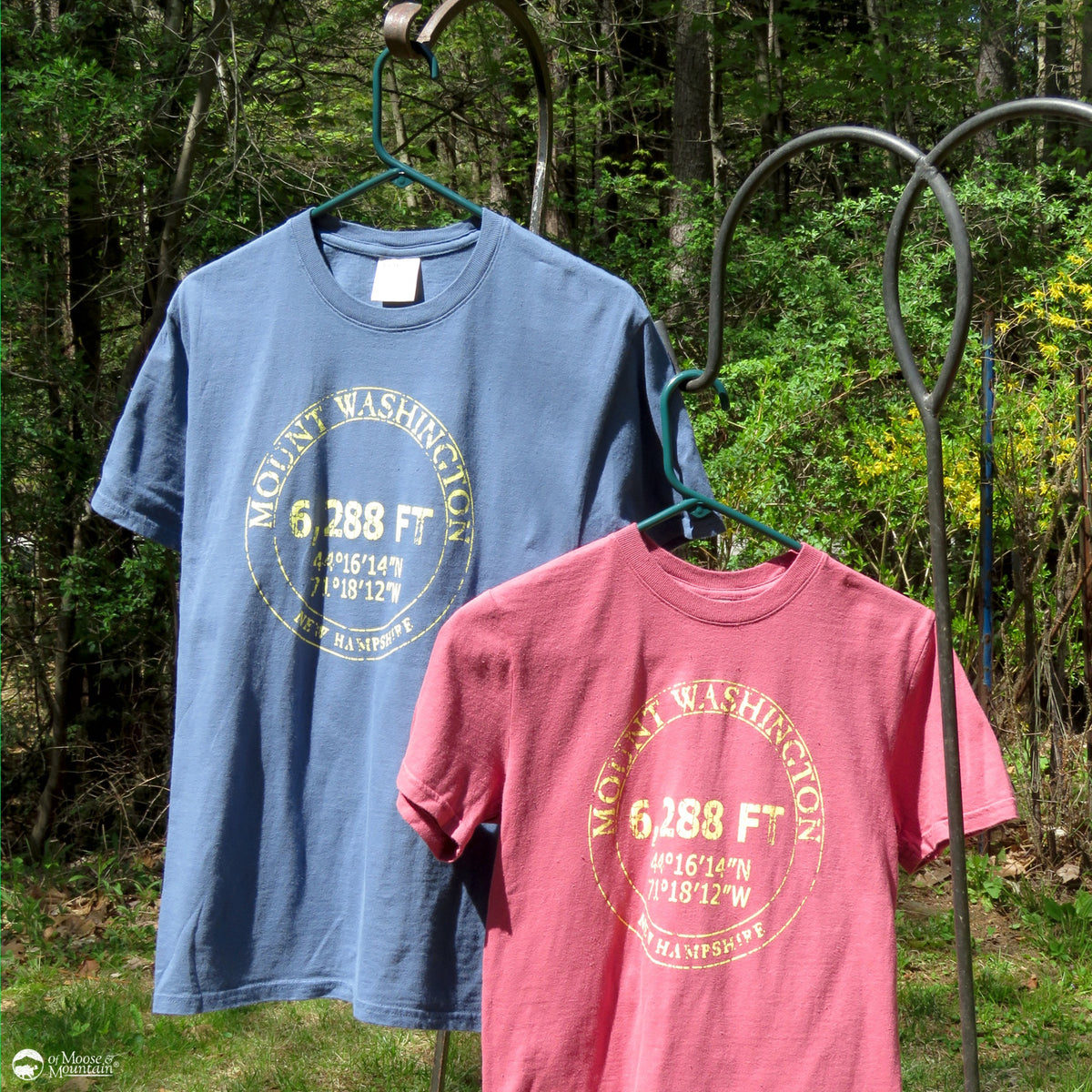Two t-shirts, one blue and one pink, with Mount Washington coordinates printed on them, hanging outdoors against a forest background.